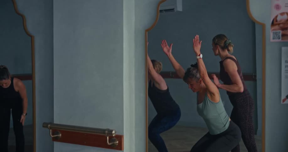 A pilates instructor helps a student with her chair pose during a group class, with their reflections visible in the studio mirrors
