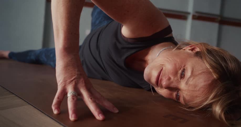 A mature woman in a yoga class holds a deep twisting stretch on her mat, breathing through the pose