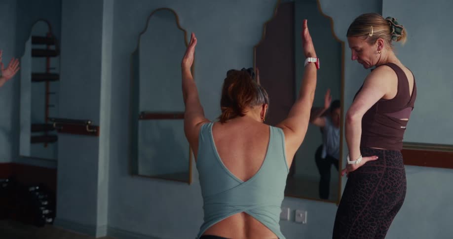 A female yoga instructor carefully corrects her student's posture and alignment during a chair pose in a quiet studio