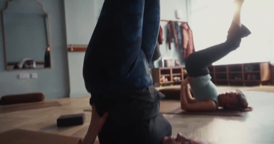 Slow upward pan following a woman as she moves into a supported shoulder stand in a tranquil yoga studio