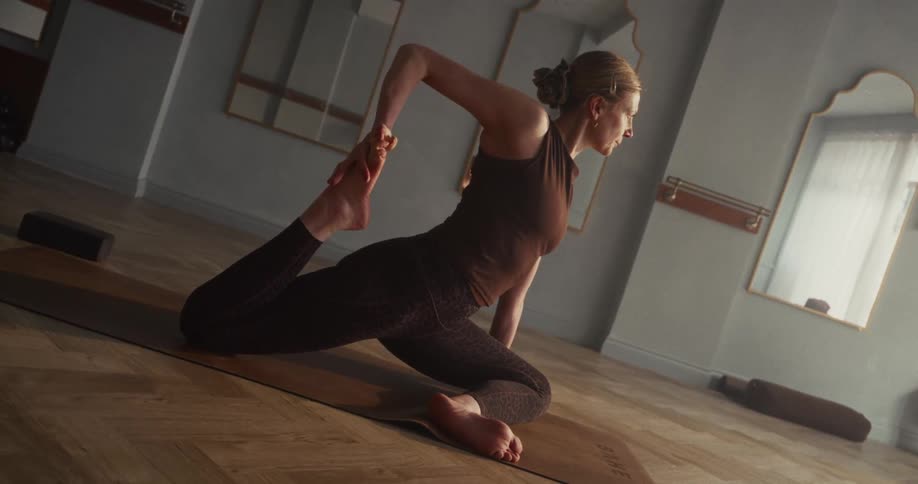 A focused woman holds a deep stretching yoga pose on a mat in a serene, sunlit studio