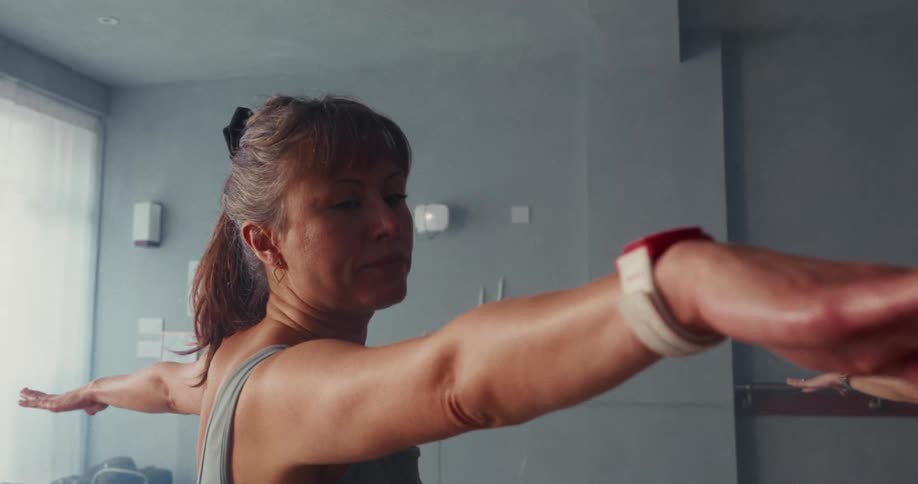 A focused middle-aged woman holds a steady warrior two pose during a yoga class in a sunlit studio
