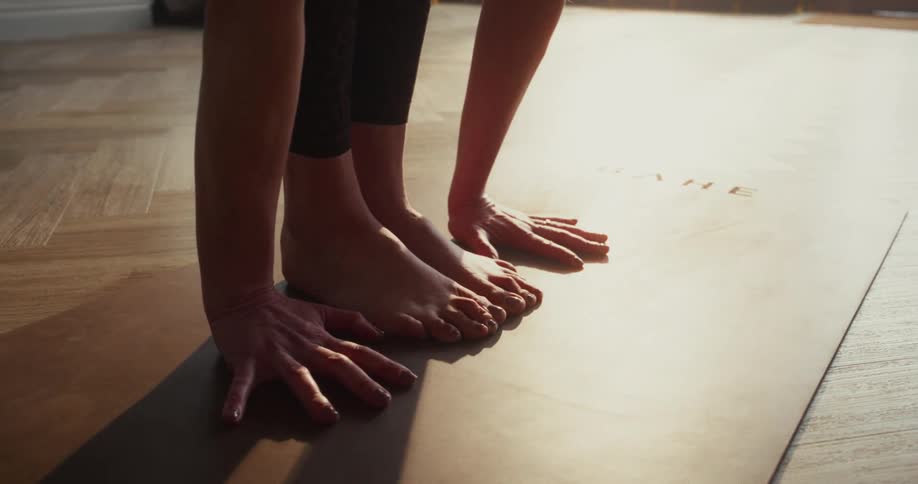 A woman performs a standing forward bend yoga pose, with a close-up on her hands and feet on a mat bathed in sunlight