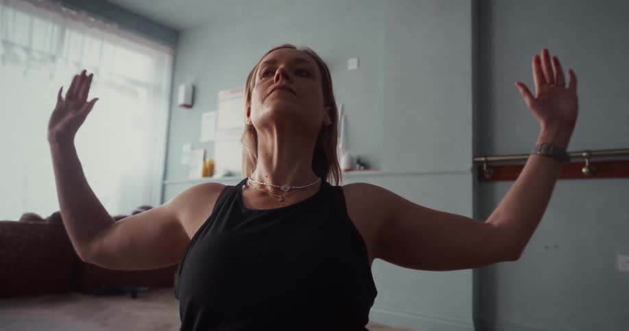 A woman in a sunlit room gracefully moves through a slow-motion yoga flow, finding her center and peace
