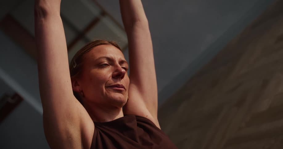 A focused middle-aged woman holds a deep stretch with her arms raised during a calm yoga session