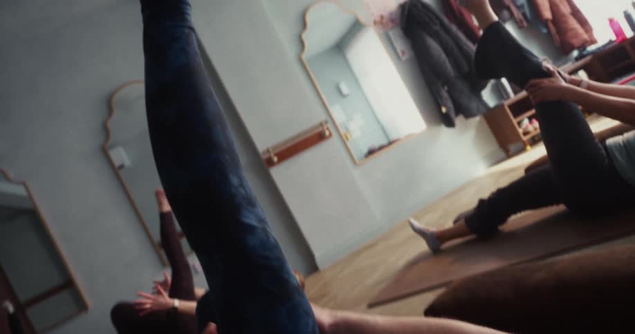 A woman in a yoga class stretches her leg towards the ceiling while lying on her back, with the camera following the movement