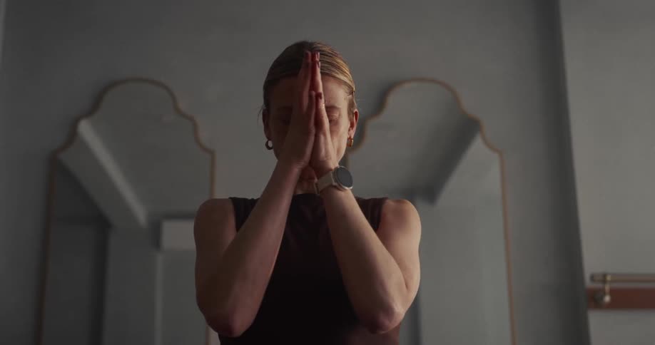 A serene woman meditates with her eyes closed and hands together in a prayer pose inside a dimly lit room
