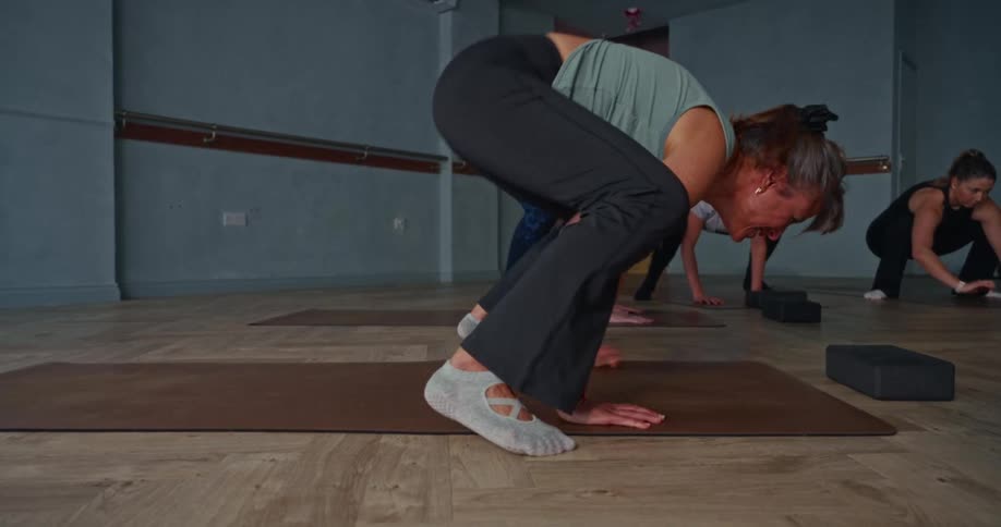 A woman in a yoga class attempts a challenging crow pose in slow motion and loses her balance
