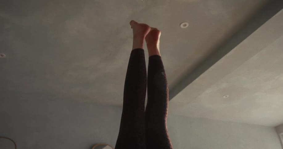 A woman gracefully transitions into a supported headstand yoga pose on a mat in a bright, sunlit studio