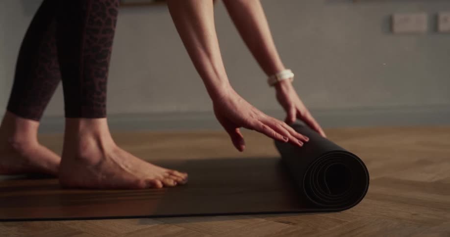 Slow motion close up of a woman's hands carefully rolling up a black yoga mat on a wooden floor after her practice