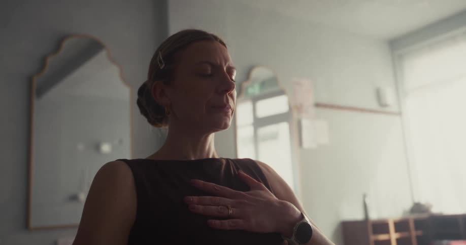 A calm woman with her eyes closed meditates with her hand on her chest in a softly lit room