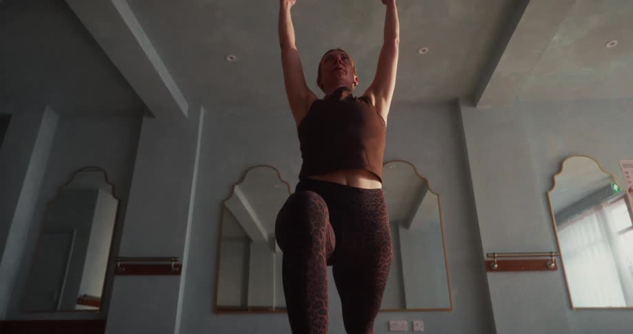 A woman in a sunlit studio gracefully moves into a high lunge yoga pose in slow motion