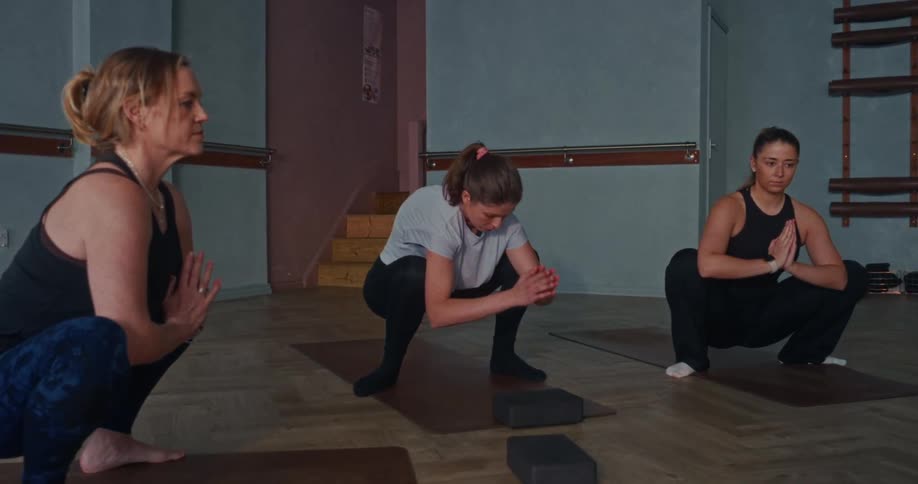 Slow zoom on a yoga instructor leading a class of women in a deep squatting pose inside a studio