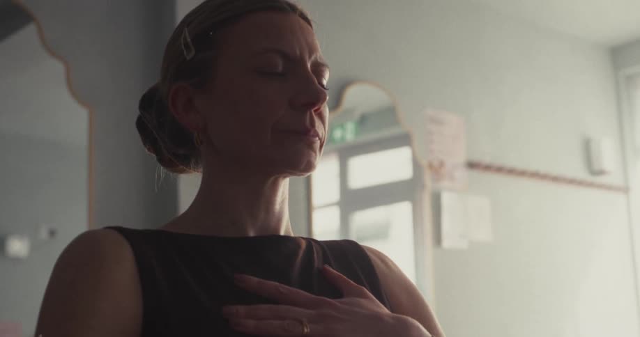 A woman with eyes closed and a hand on her heart practices a calming breathing exercise in a softly lit room