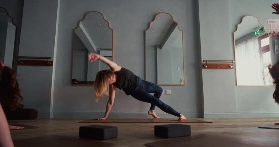 A woman gracefully transitions from a downward-facing dog into a wild thing yoga pose during a class in a serene studio, filmed in slow motion