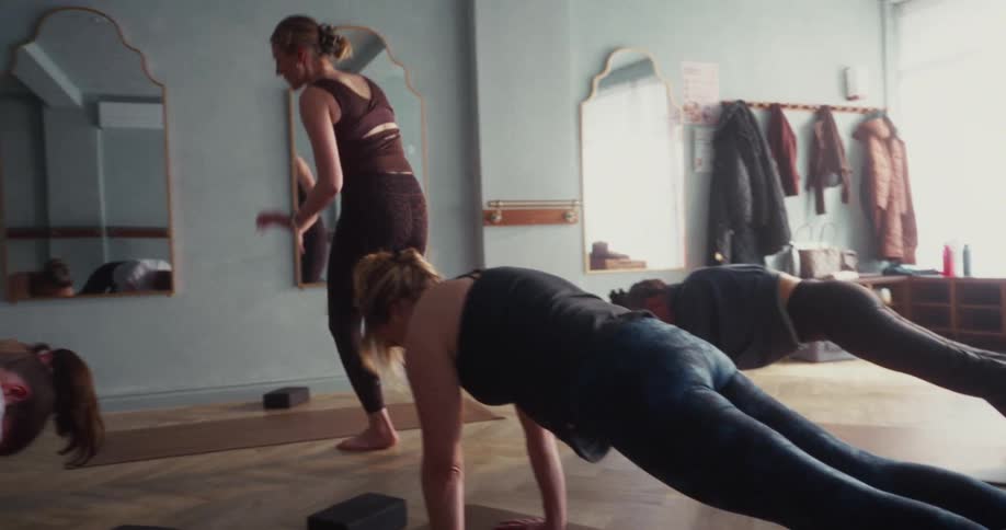 An instructor guides her class through plank poses, offering gentle corrections in a sun-drenched yoga studio in slow motion