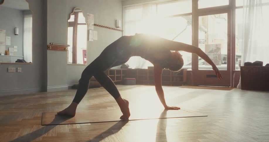 A flexible woman holds a deep backbend yoga pose on her mat in a beautiful, sun-drenched studio