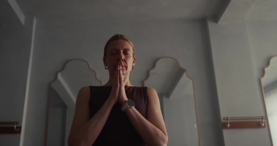 A calm woman with her eyes closed meditates indoors with her hands in a prayer pose