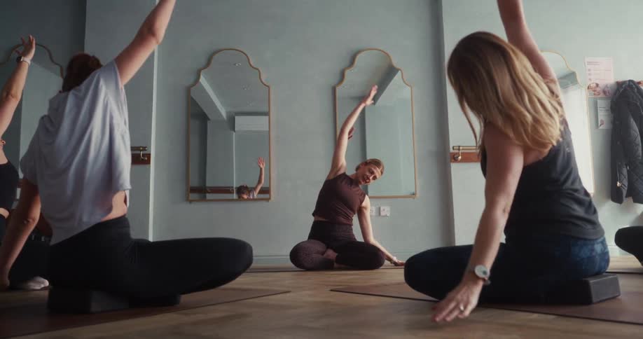 Women follow an instructor through calming, seated stretches during a peaceful yoga class in a well-lit studio