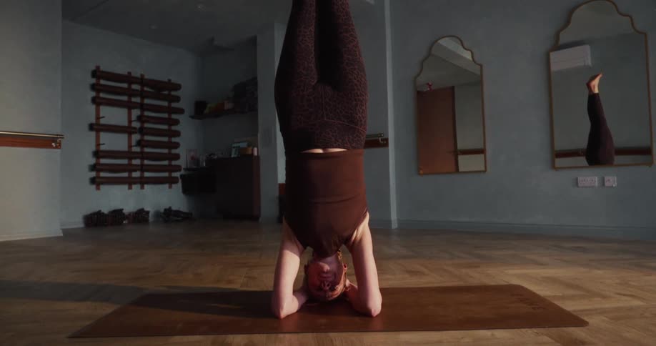 A focused woman holds a steady headstand yoga pose on a mat in a quiet studio, revealed in a slow pullback shot