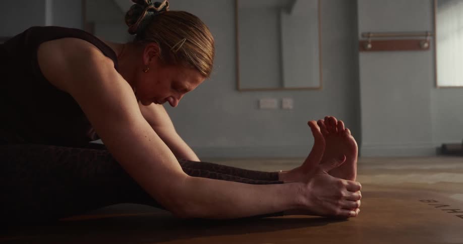 A focused woman in a calm studio setting gracefully performs a seated forward bend yoga stretch in slow motion