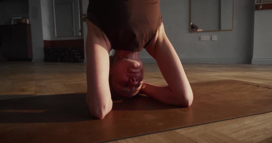 A woman gracefully performs a supported headstand yoga pose on a mat in a sunlit studio