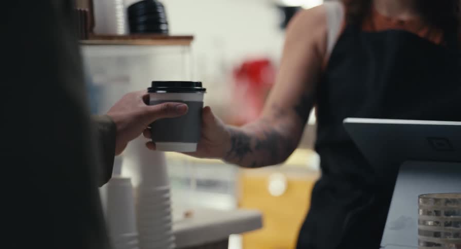 A tattooed barista hands a steaming hot coffee in a paper cup to a customer at the cafe counter