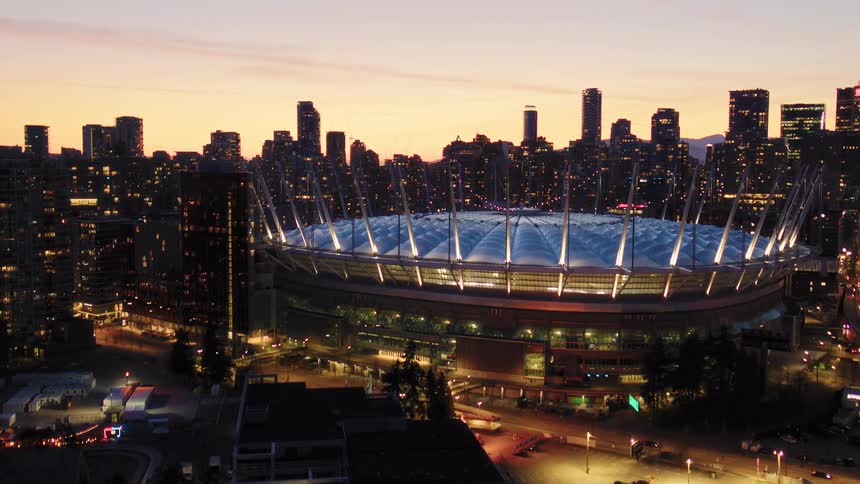A cinematic aerial shot tilts up from the water's reflection to reveal the illuminated BC Place stadium and the Vancouver city skyline at sunset