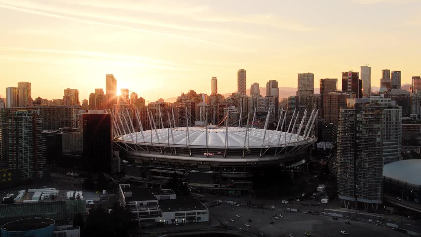 Stunning aerial view pulling back from BC Place stadium, revealing the expansive Vancouver city skyline as the sun sets with a golden glow