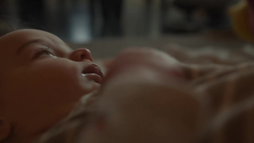 An intimate shallow-focus close-up of an infant lying on its back and looking up curiously in a dimly lit room