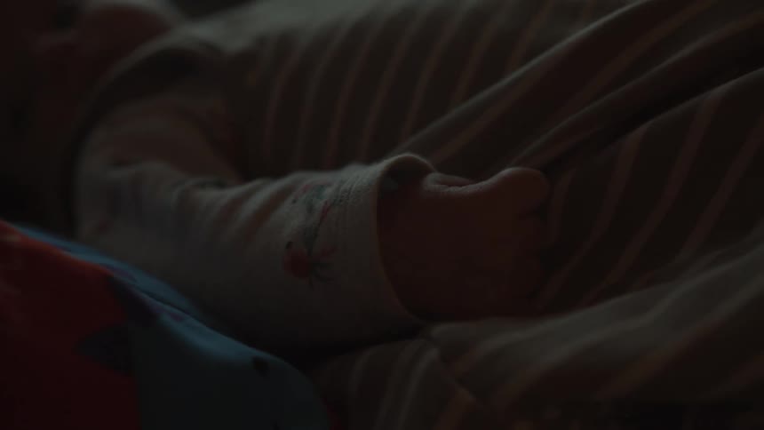 A tender close-up focuses on the small hand of a sleeping baby who is resting peacefully in a dimly lit room