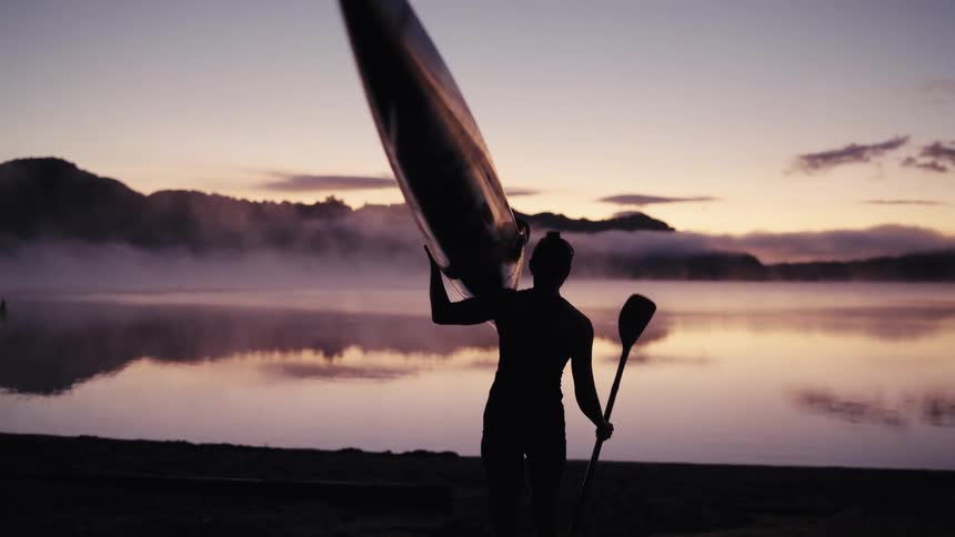 A silhouetted woman carries her kayak on her shoulder as she walks toward a calm, misty lake at sunrise