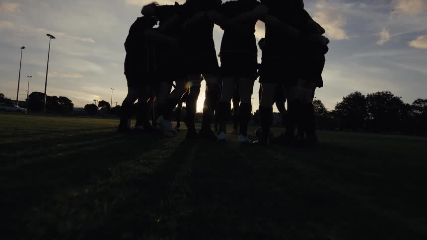 A team of male athletes in silhouette form a motivational huddle on the field as the sun sets behind them