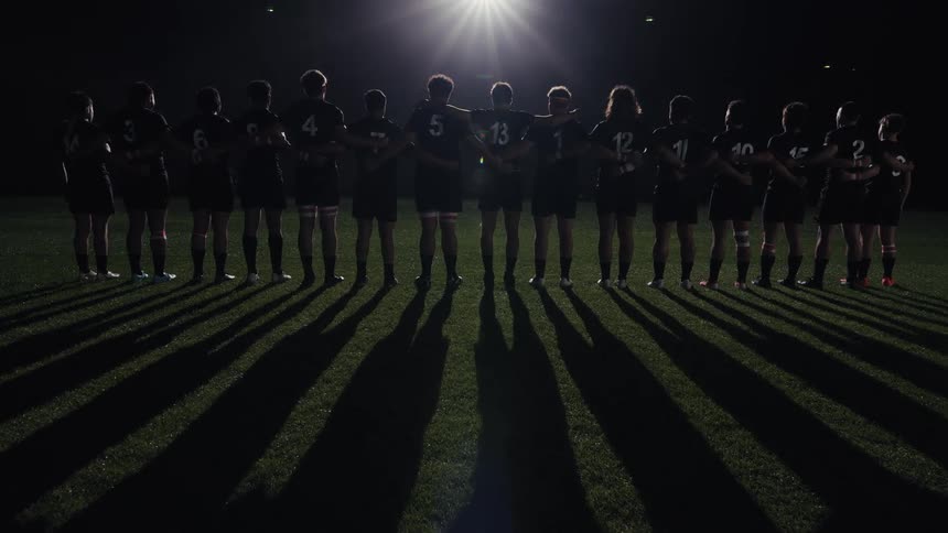 A silhouetted rugby team stands united arm-in-arm on a field at night, facing a bright spotlight that casts long, dramatic shadows