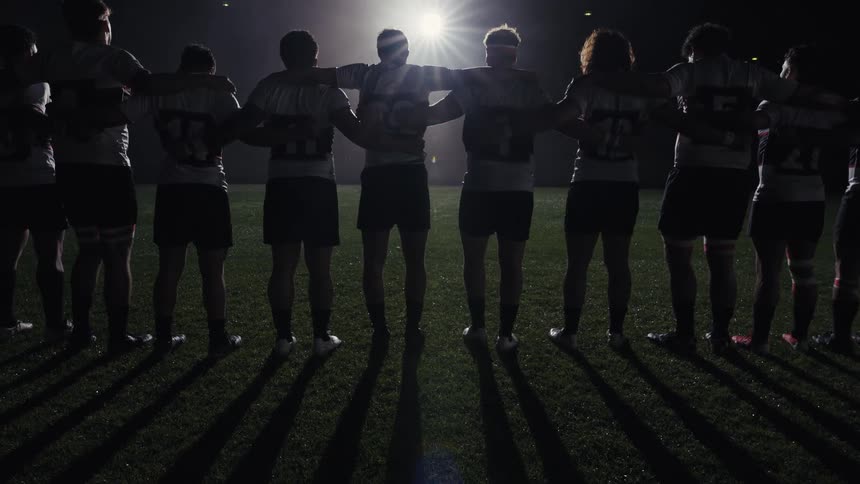 A rugby team stands united on the field at night, silhouetted by a bright stadium light as they put their arms around each other in a show of teamwork