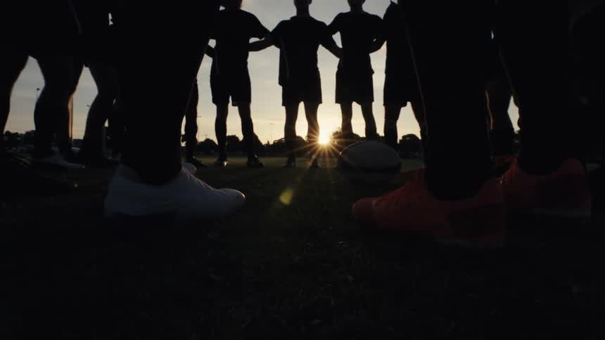 A low-angle shot shows a silhouetted sports team forming a motivational circle on a grassy field as the sun sets behind them