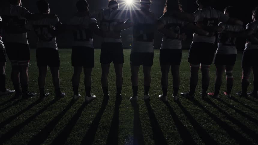 A silhouetted rugby team unites on the field, their arms around each other under a dramatic stadium light at night