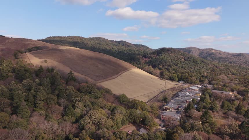A slow aerial pullback reveals the scenic Mount Wakakusa in Nara, Japan, with its dry grassy slopes and a nearby village under a blue sky