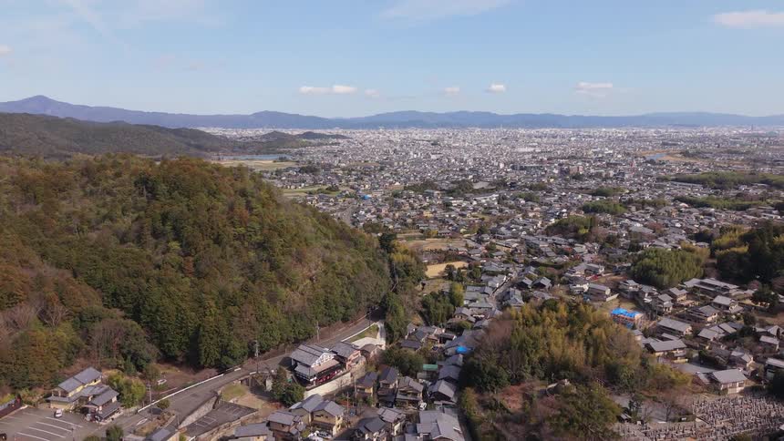 An expansive aerial view reveals a Japanese city spreading out from the base of a forested mountain on a sunny day