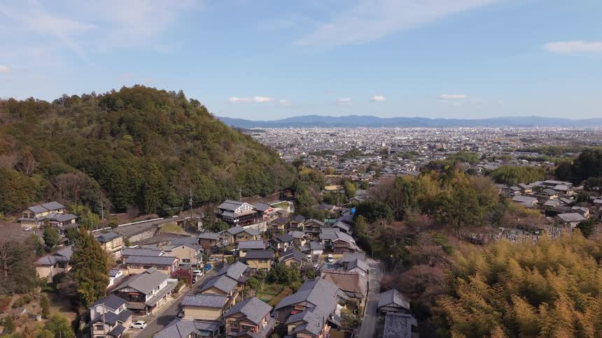 Aerial drone footage ascends over a historic village in the Arashiyama district of Kyoto, Japan, contrasting the traditional town with the modern cityscape beyond