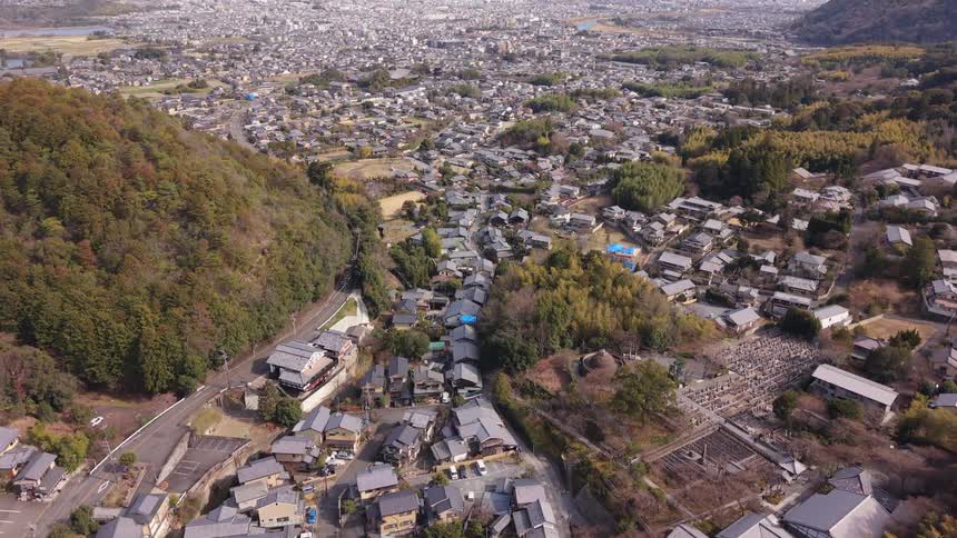 Slowly rising aerial view reveals the sprawling cityscape of Nara, Japan, beyond a traditional hillside village