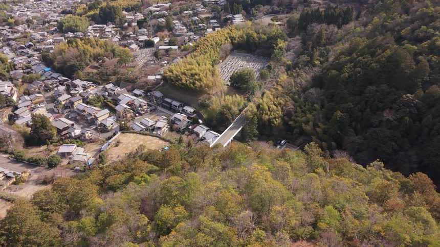 Aerial pullback reveals a quiet rural Japanese village nestled in a green valley next to a dense forest and cemetery