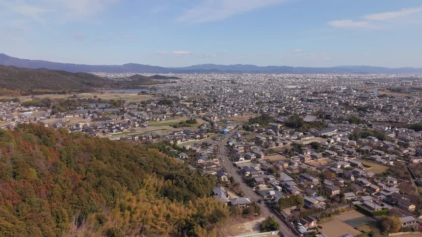 Aerial view flying over an autumn-colored forest hill towards a sprawling Japanese city on a clear day