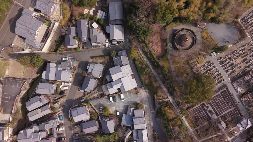 A slow ascending aerial top-down view reveals a quiet Japanese village with traditional architecture, nestled beside a bamboo forest and an ancient cemetery
