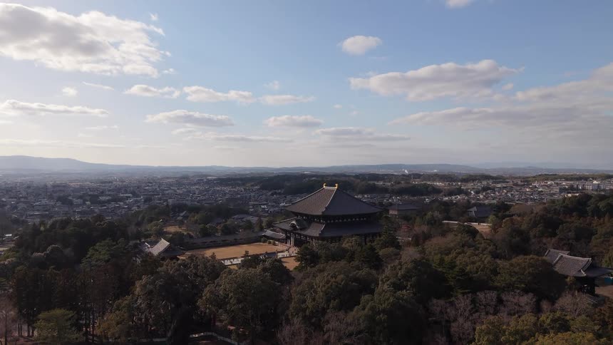 An expansive aerial view reveals the majestic Todai-ji Temple nestled in a dense forest, with the city of Nara sprawling towards distant mountains under a blue sky