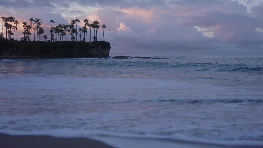 Slow motion waves wash ashore at dusk with a silhouetted cliff of palm trees against a dramatic sky