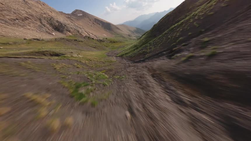 An exhilarating FPV drone flight surfs the rugged terrain of a mountain valley, skimming over scree slopes and grassy hillsides