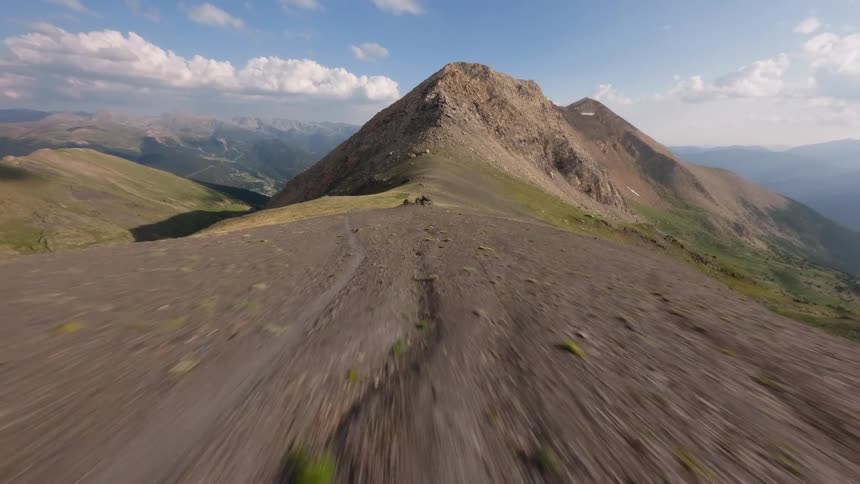 A thrilling first-person view drone flight speeds low along a rocky and grassy mountain ridge towards a distant peak