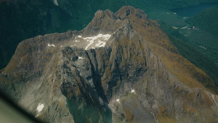 A stunning aerial view from an airplane captures a rugged, snow-dusted mountain peak towering over a vast, green forested valley