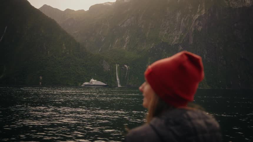 A woman in a red beanie watches a ferry sail past a majestic waterfall in the Norwegian fjords